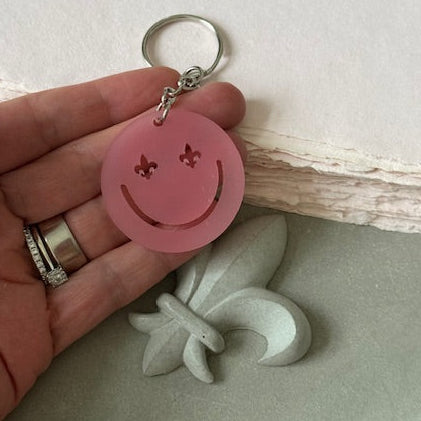 „Runder Schlüsselanhänger aus roséfarbenem Acryl mit Smiley-Design und Lilien-Augen, symbolisiert Eleganz und Lebensfreude, fotografiert auf einem hellen Untergrund.